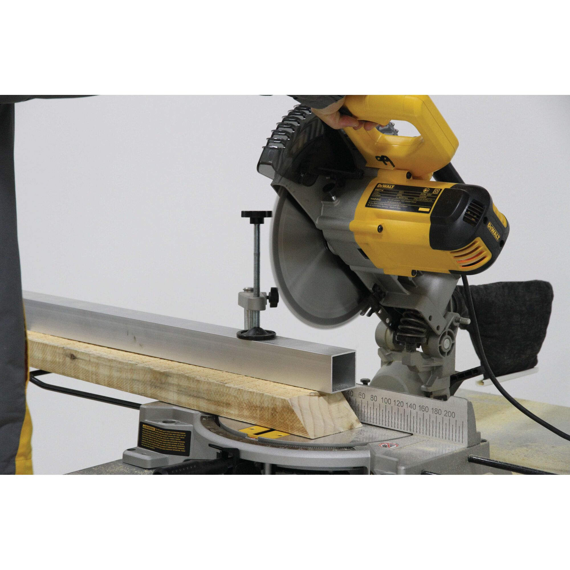 A DEWALT DW714 miter saw being used to cut a piece of metal and a wooden beam. The saw is yellow and black, and the metal and wood are secured on the saw base.
