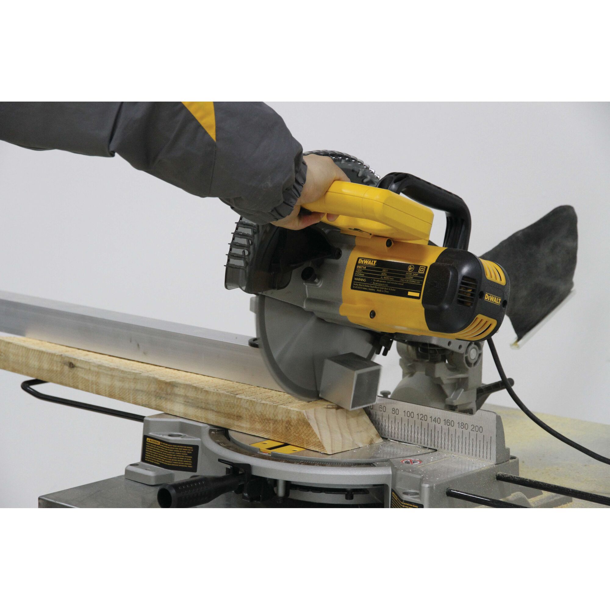 A person operating a DEWALT DW714 miter saw is cutting a piece of wood at an angle. The saw is yellow and black and is placed on a workbench.