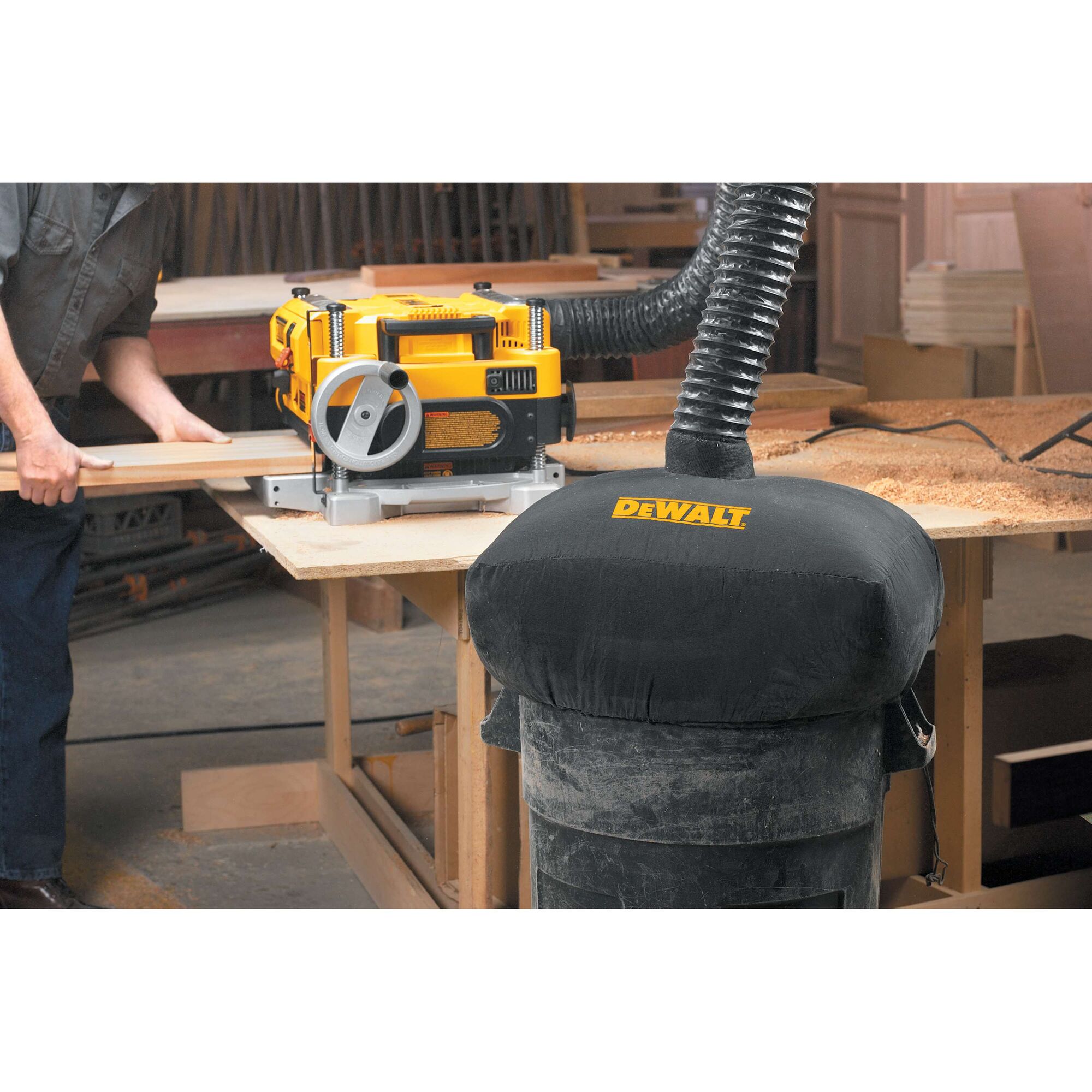 A yellow DEWALT wood planer sits on a workbench, with a dust collection system attached to it. Sawdust is scattered on the bench, and a person is using the planer in a workshop setting. The dust collector has a black cover with the DEWALT logo.