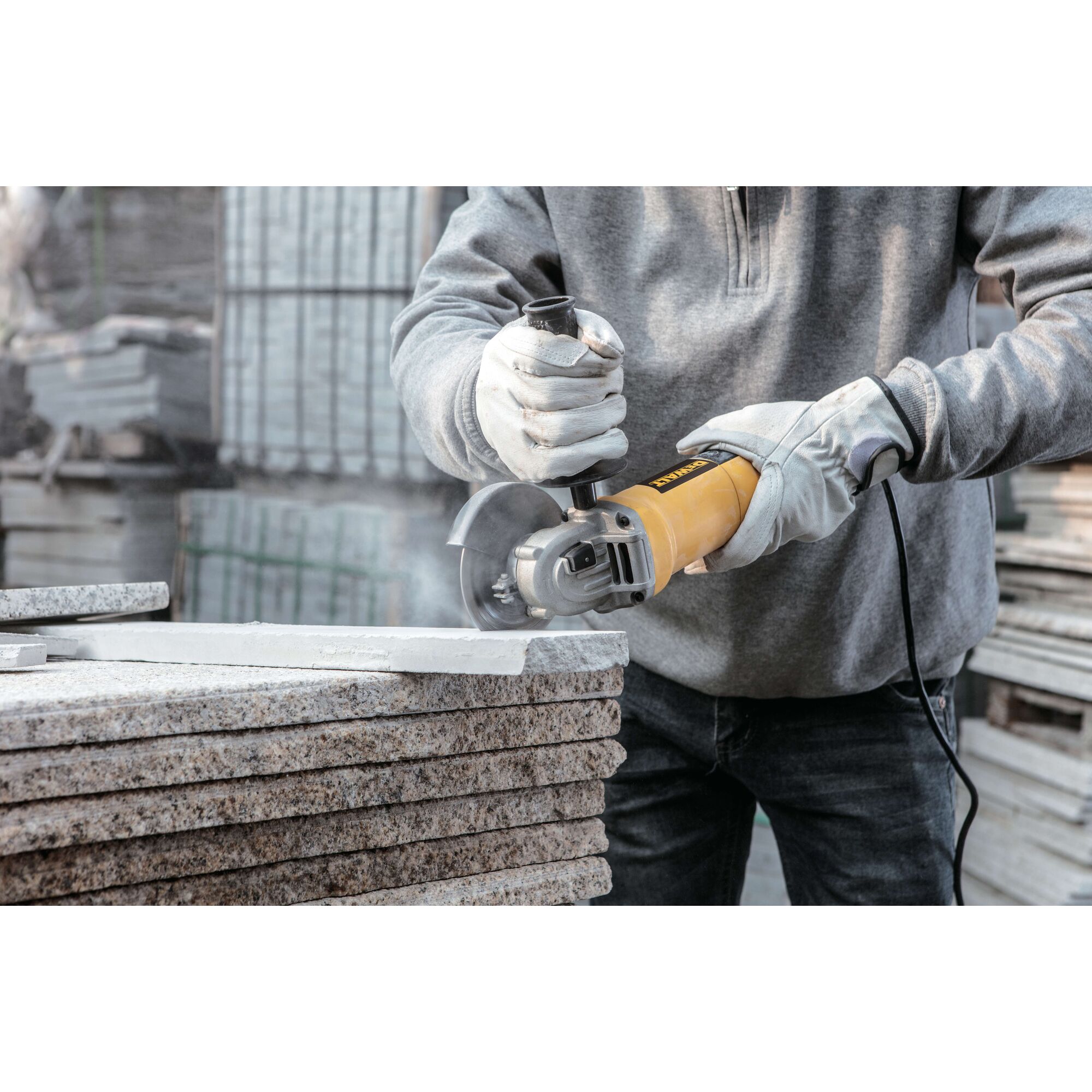 Person wearing gloves using a yellow DEWALT angle grinder to cut a stone slab outdoors, with dust visible around the cutting area.