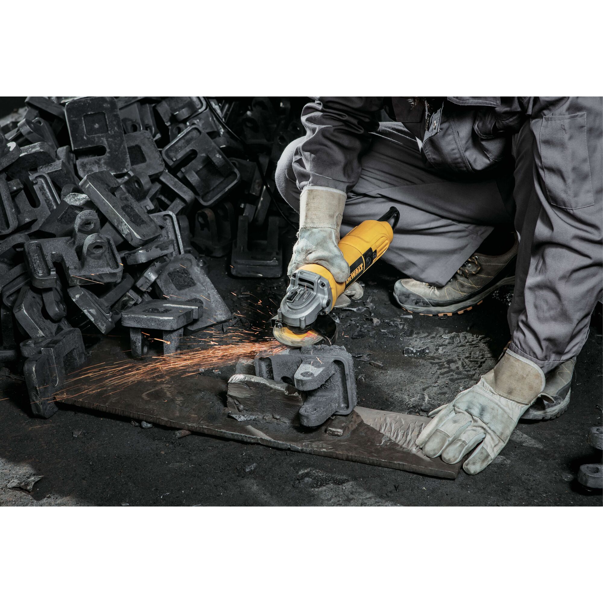 A person wearing protective gloves and work clothes is using a yellow DEWALT angle grinder to grind a piece of metal, producing sparks. There are several metal parts scattered on the floor around the person.