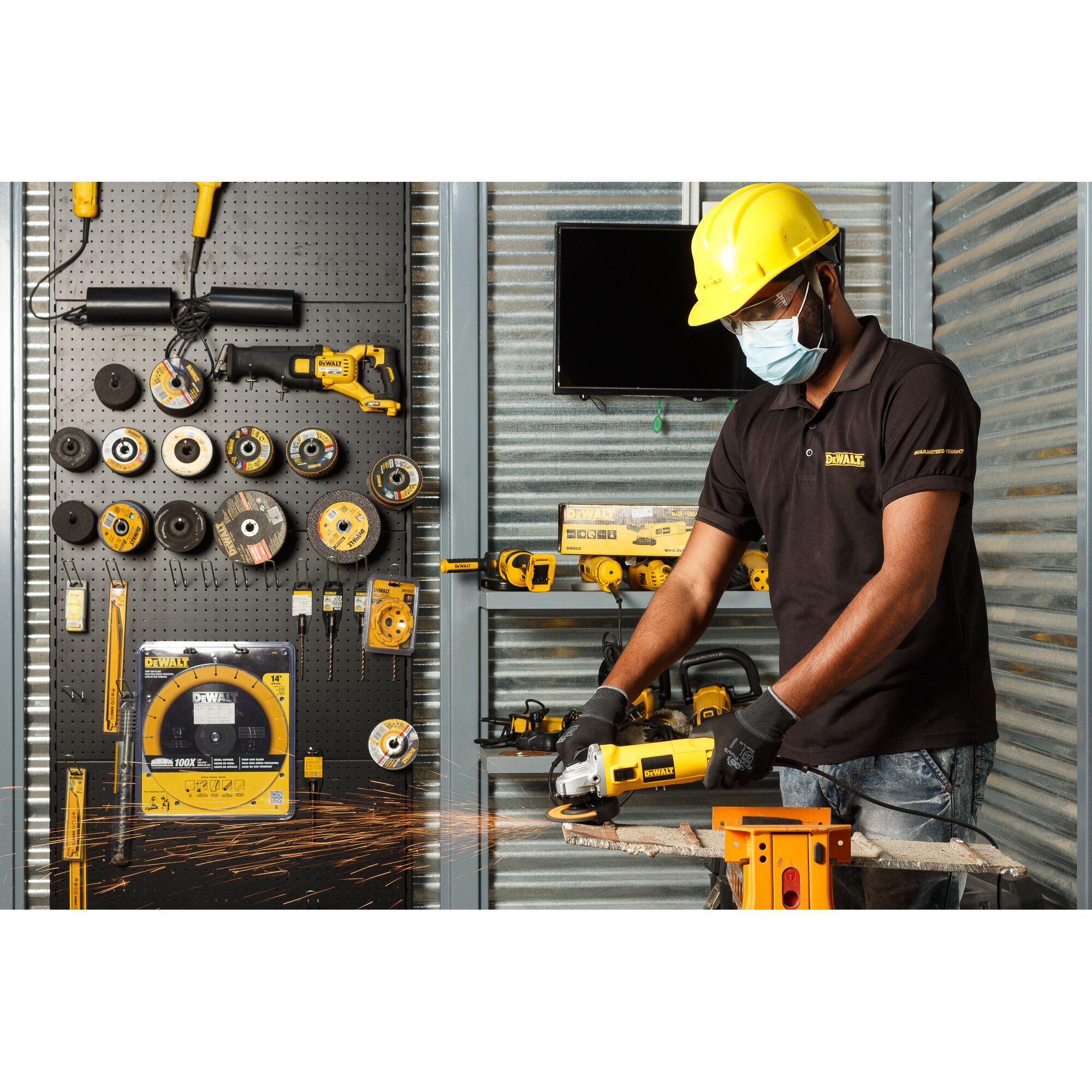 A person wearing a yellow hard hat and gloves is using a DEWALT DW802 power tool to cut metal, producing sparks. The workspace has a pegboard displaying various DEWALT accessories and tools, and a metal wall in the background.
