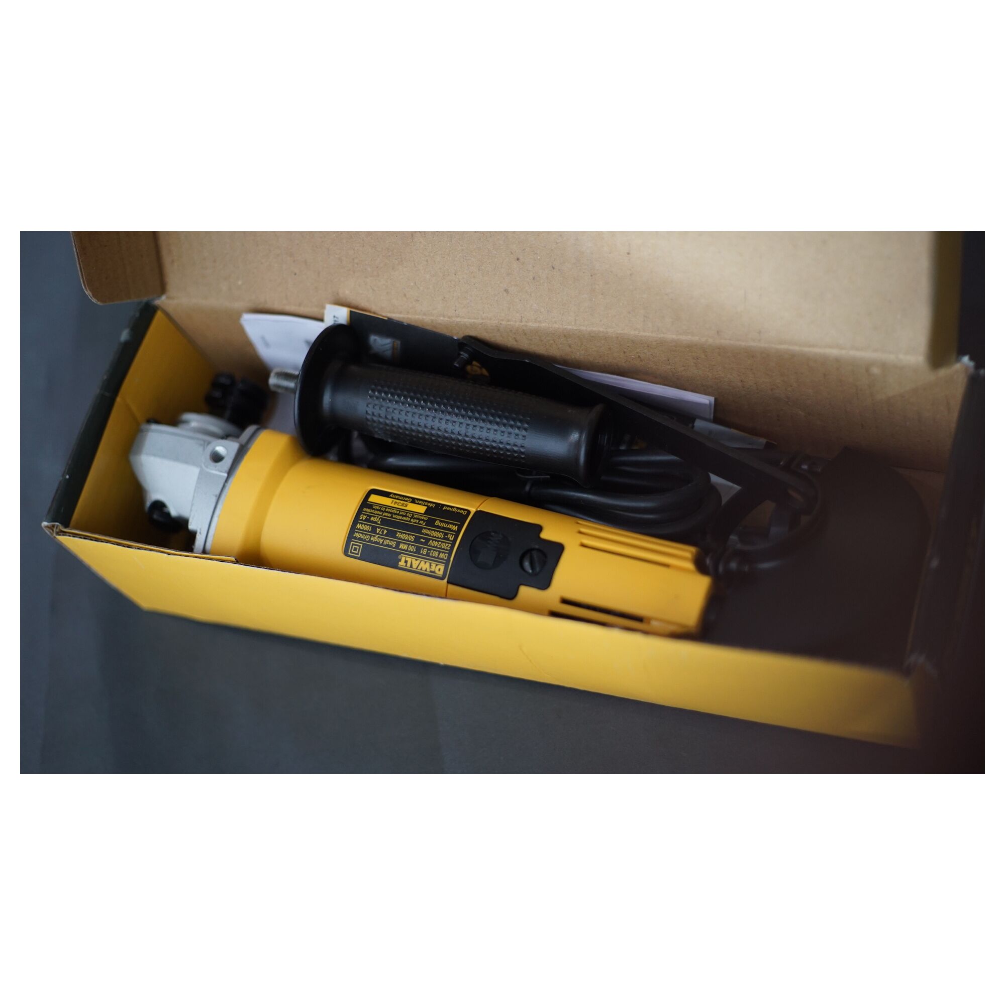 A DEWALT DW803 angle grinder in yellow and black, shown inside its open cardboard packaging with a side handle and power cord visible.