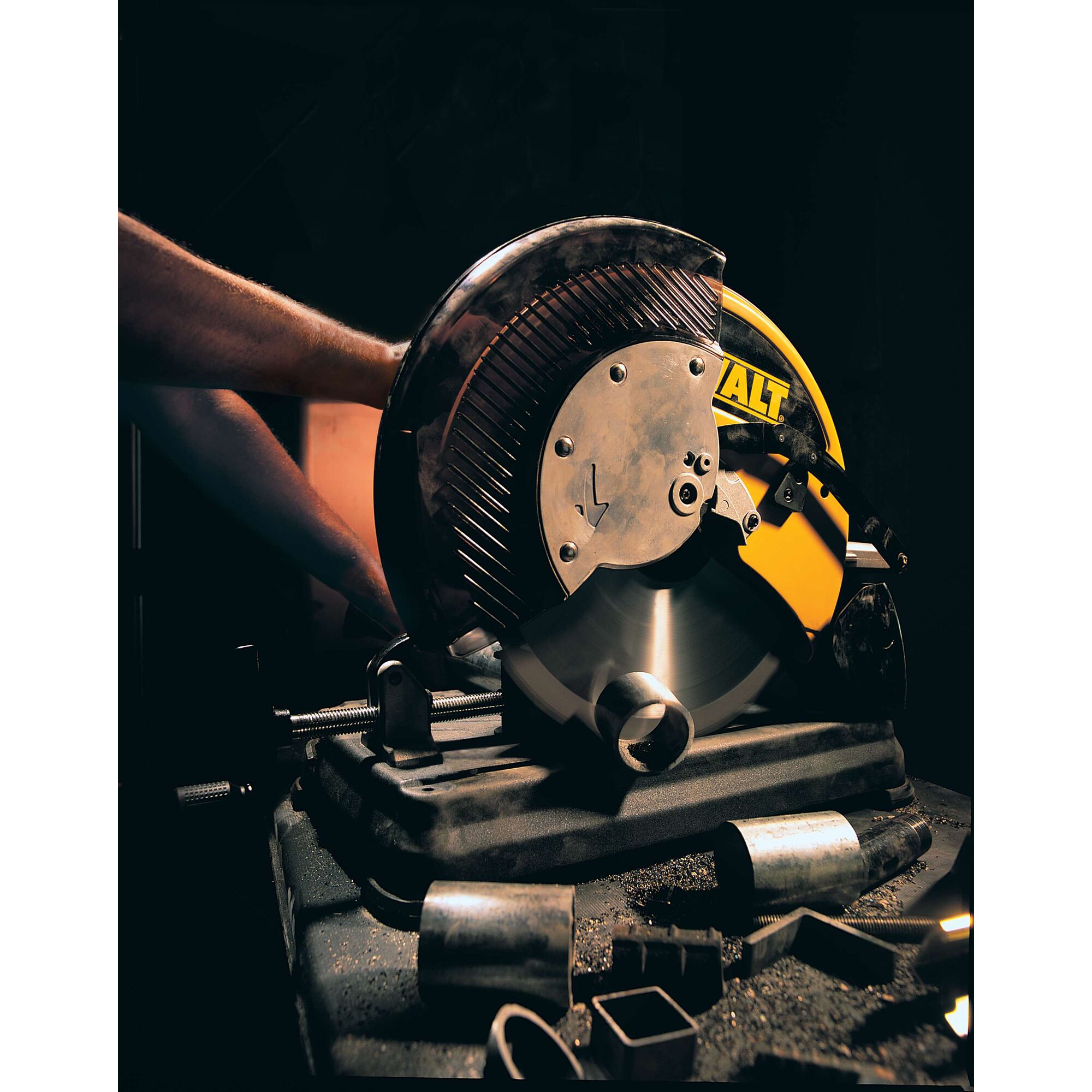 A DEWALT metal cutting saw is in use, cutting through a cylindrical metal pipe. The saw has a large circular blade and is mounted on a sturdy base. There are several pieces of cut metal and metal shavings scattered on the table around the saw.