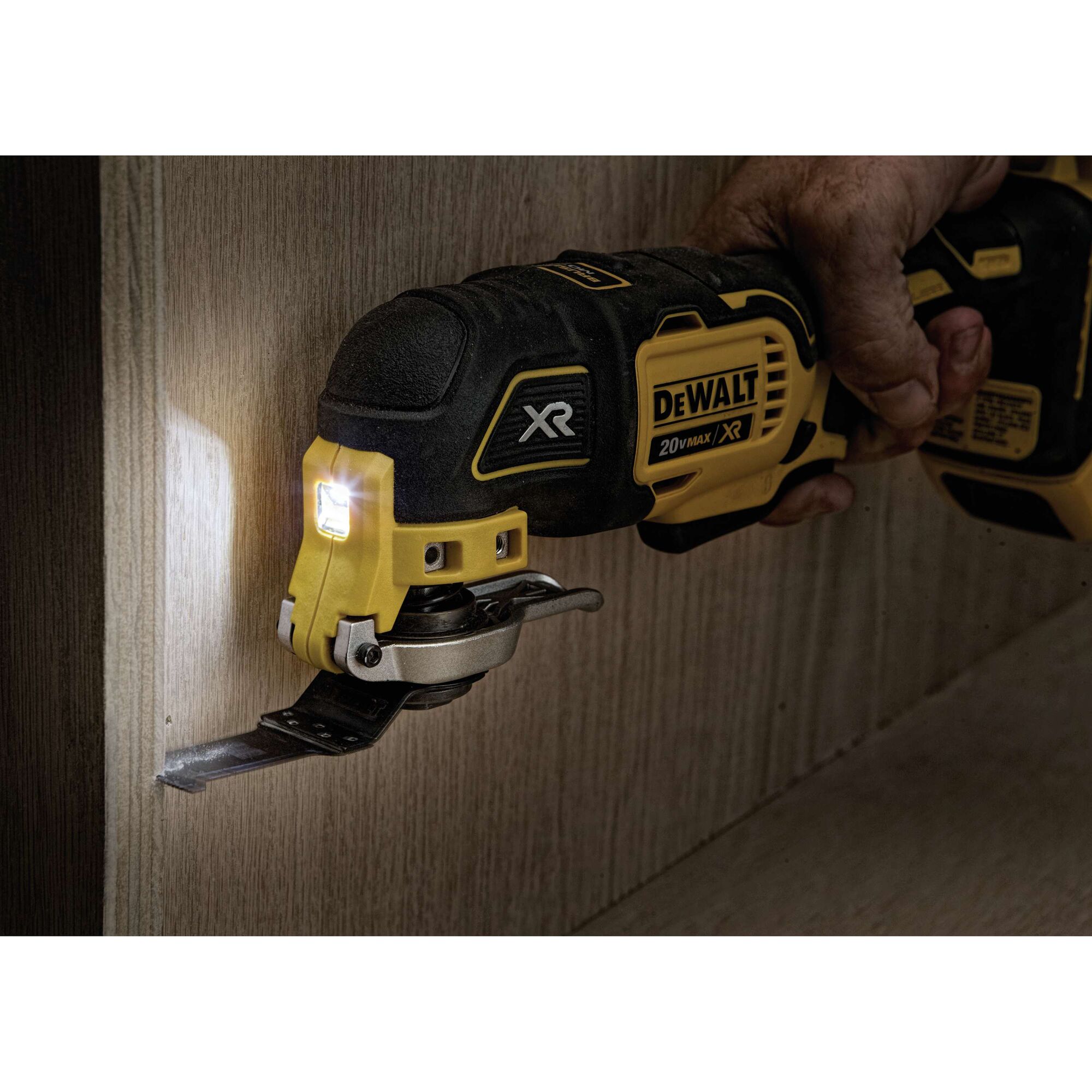 A DEWALT XR 20V Max oscillating multi-tool is being used to cut into a wooden surface. The tool's LED light is illuminated, and a person is holding the tool.