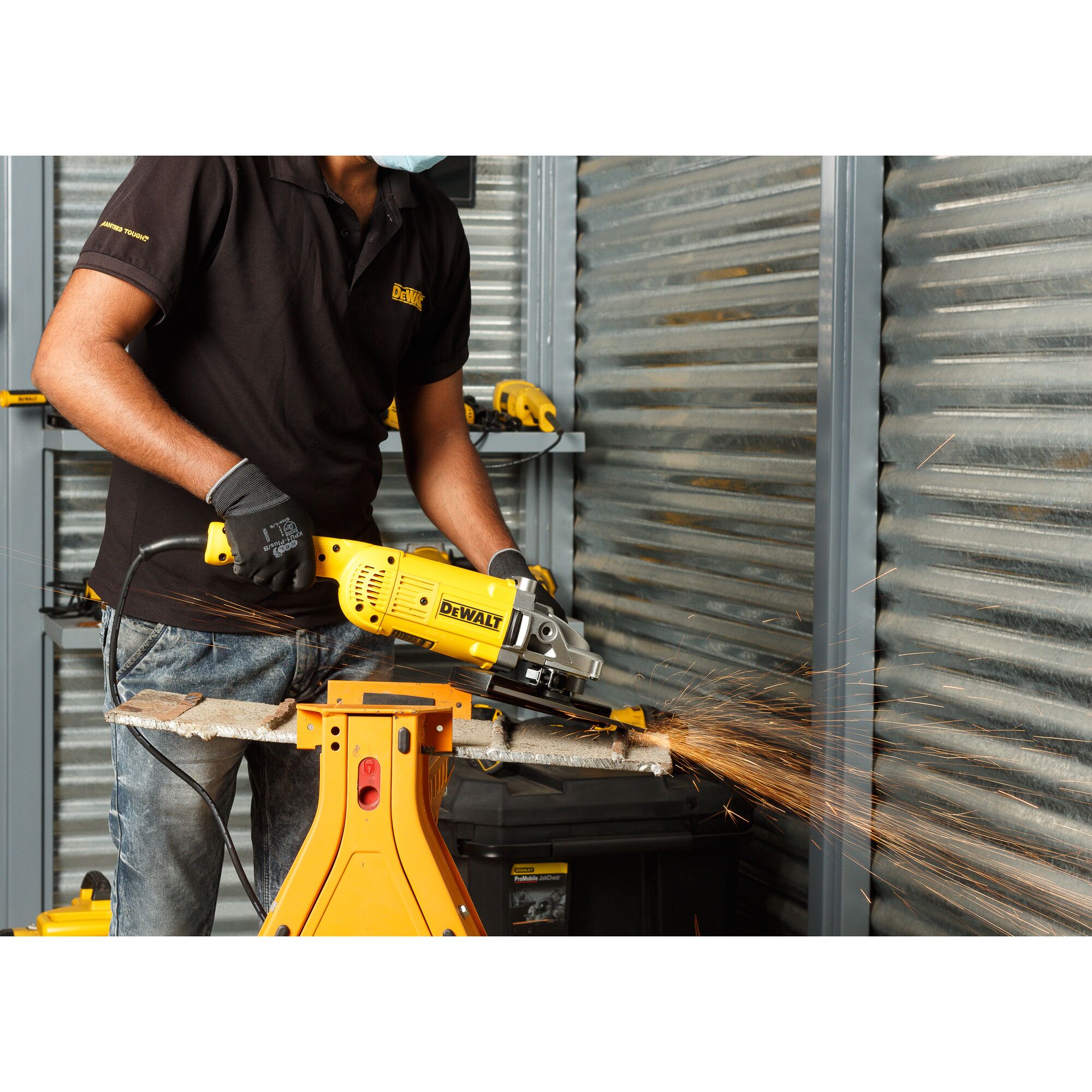 A person wearing gloves operates a yellow DEWALT DWE492 angle grinder, cutting through a metal pipe with sparks flying, inside a workshop with metal panels and other DEWALT tools visible.