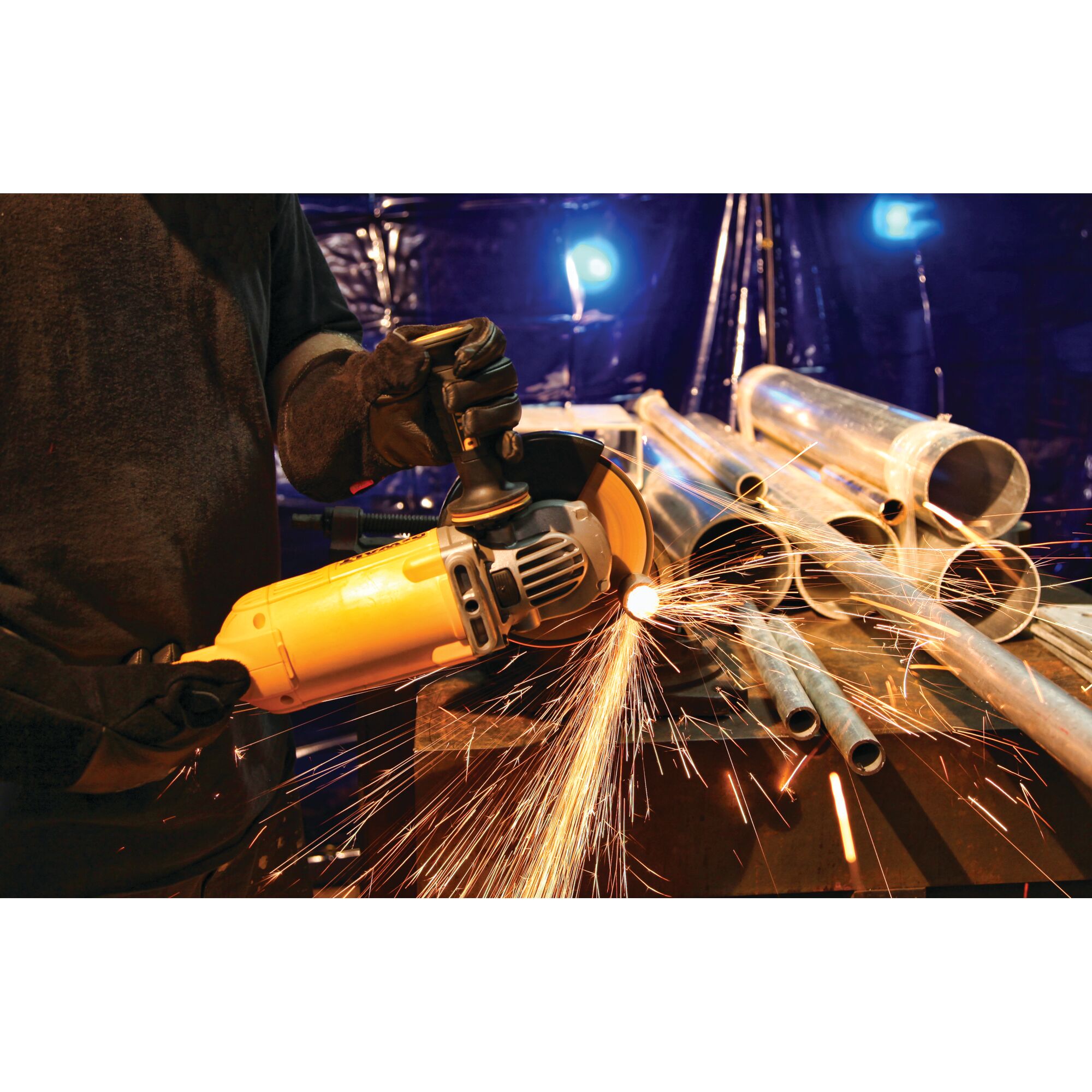 A person wearing gloves uses a yellow DEWALT angle grinder to cut metal pipes, producing visible sparks, in an industrial workshop setting.