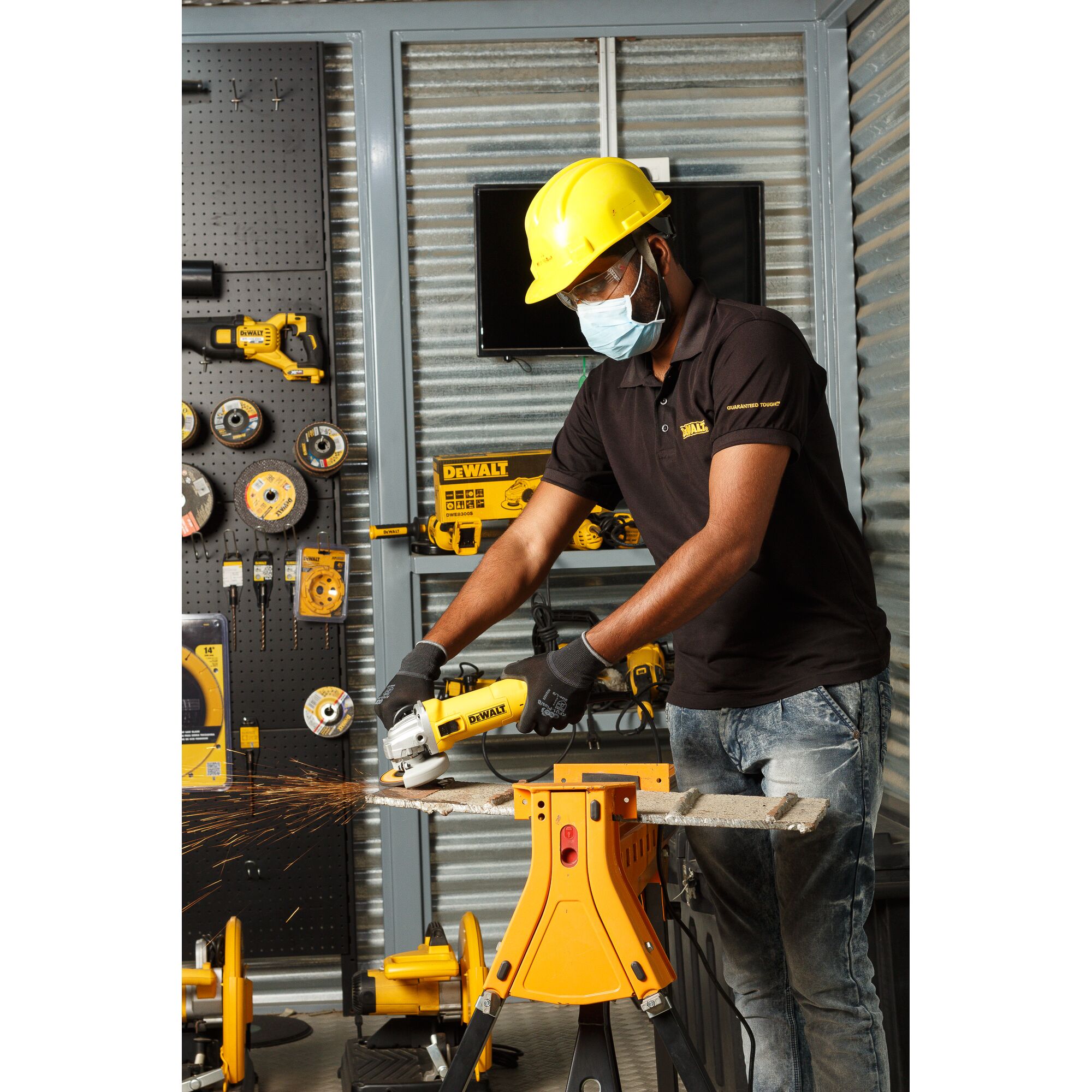 A person wearing a yellow safety helmet, face mask, and gloves using a DEWALT DWE8300S angle grinder to cut a piece of material supported on a yellow stand. The background shows various DEWALT tools and accessories in a workshop setting.