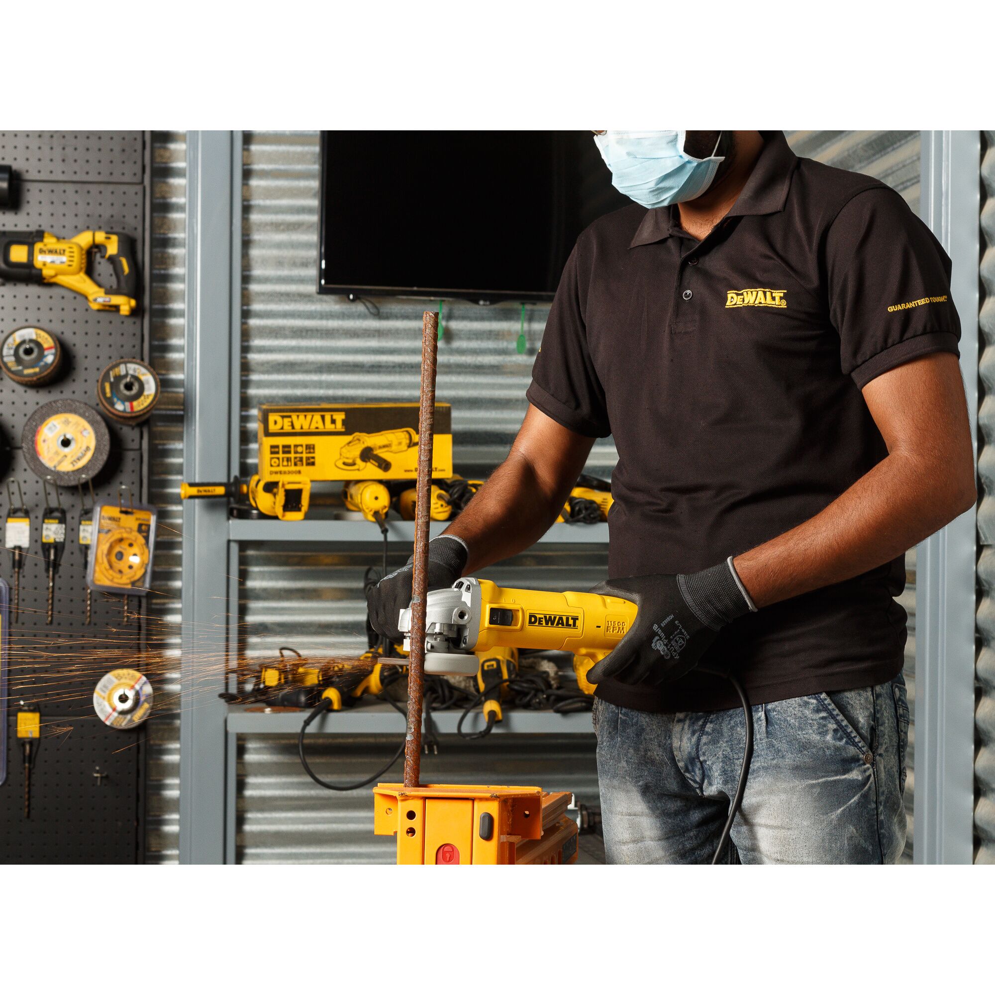 A person wearing gloves and a face mask uses a DEWALT angle grinder to cut a metal rod in a workshop. Various DEWALT tools and equipment are visible in the background.