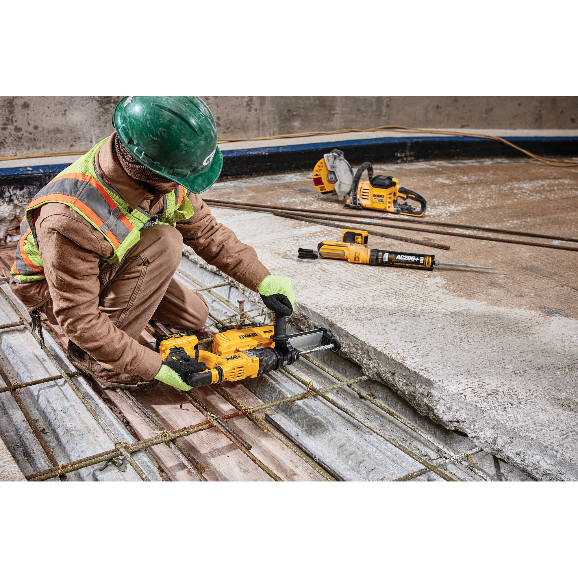 A construction worker wearing safety gear uses a yellow DEWALT power tool to drill into concrete on a worksite. Other DEWALT tools are visible on the floor nearby.