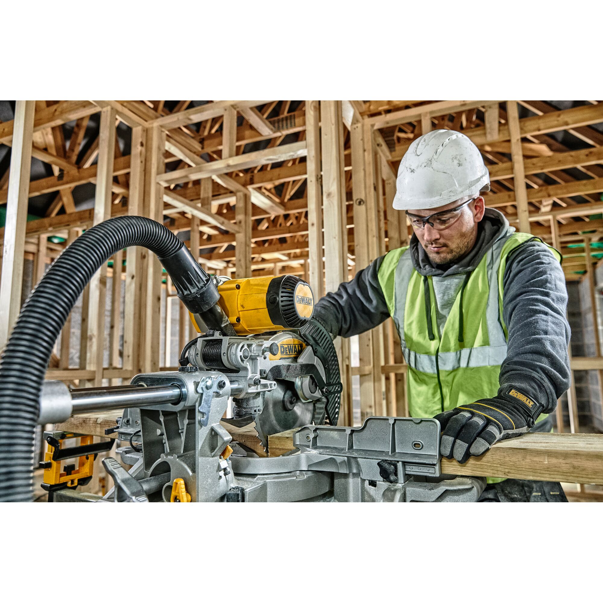 A worker wearing a safety vest and hard hat operates a DEWALT DWS727 miter saw in a wooden frame construction site. The saw is connected to a dust extraction hose.