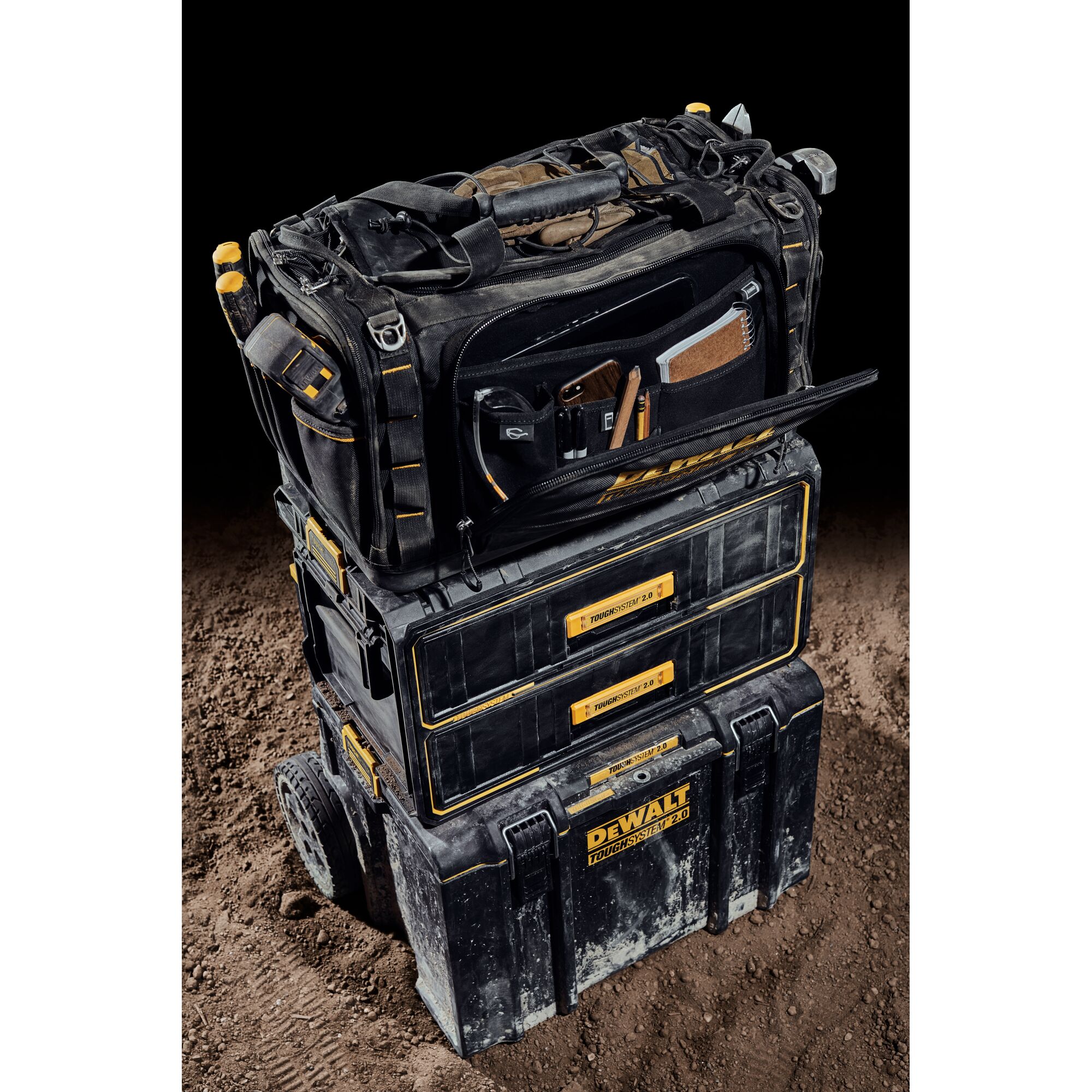 A DEWALT ToughSystem 2.0 tool storage setup is shown, consisting of three stacked black and yellow modular toolboxes. The top bag is open, revealing organized compartments with various tools and accessories. The setup is placed on a dirt surface with a black background.