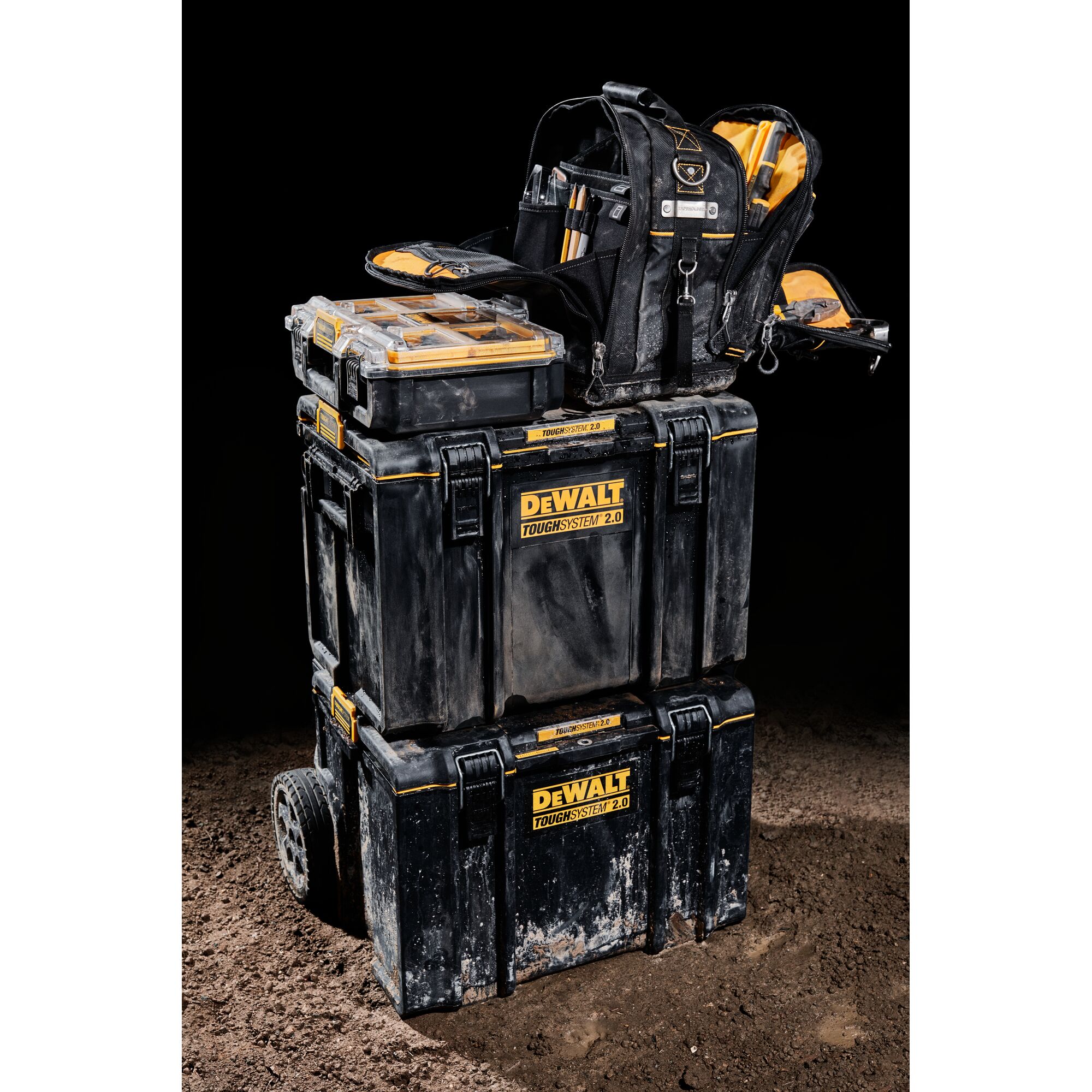 Three stacked DEWALT ToughSystem 2.0 toolboxes are shown, with a black tool backpack and a transparent organizer box on top. The toolboxes and backpack display visible dirt, suggesting use on a construction site.